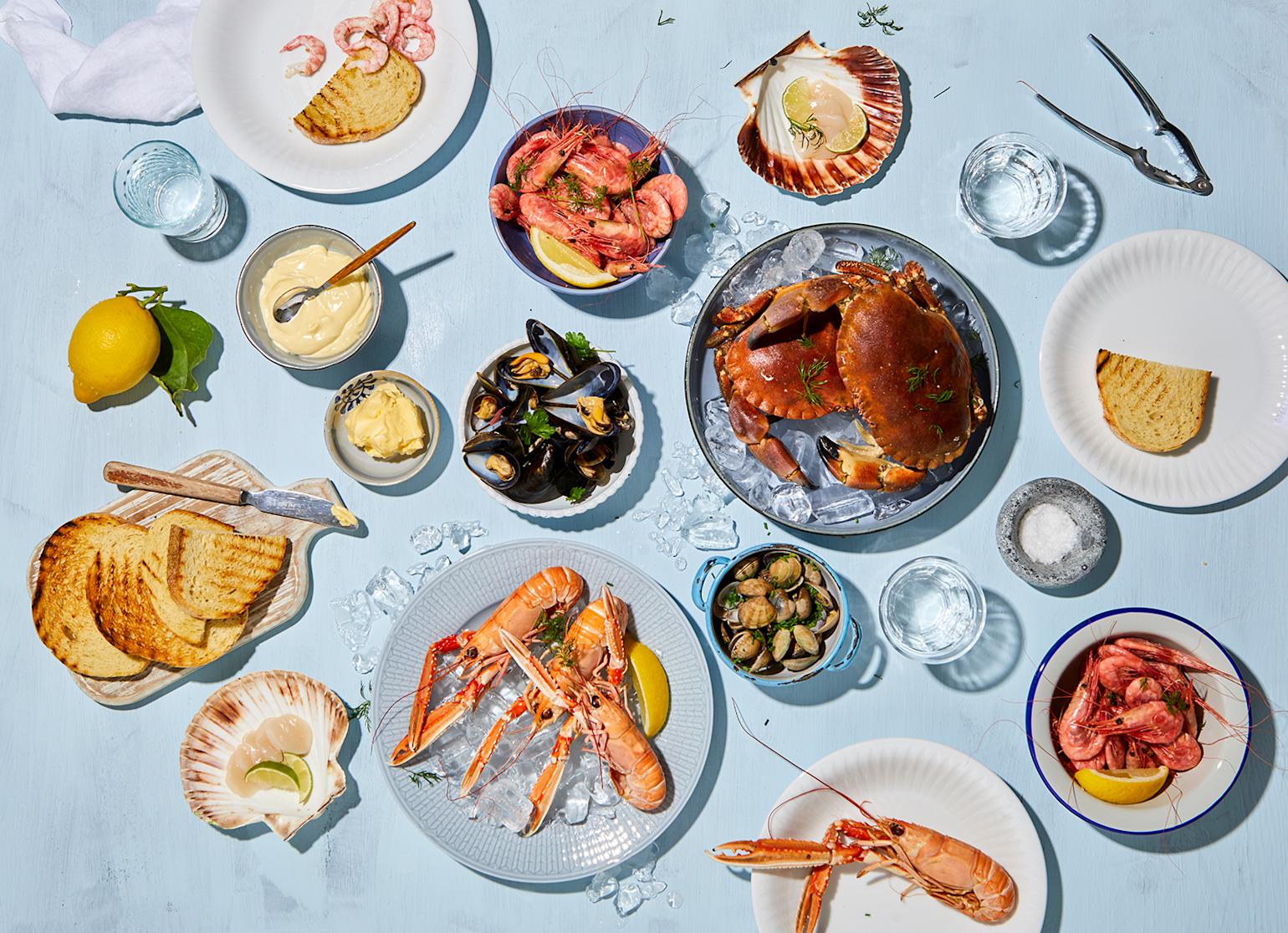 Et utvalg skalldyr på forskjellige fat og i skåler, omringet av skåler med tilbehør, ristet brød og frisk sitron.