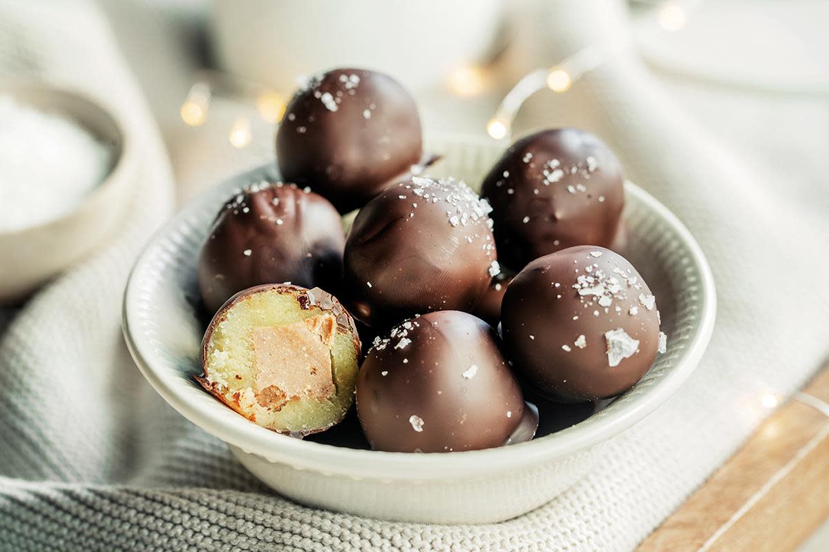 Marsipankuler fylt med nougat, trukket med mørk sjokolade, med et dryss havsalt. Servert i hvit skål. Lys stoffbakgrunn med små julelys.