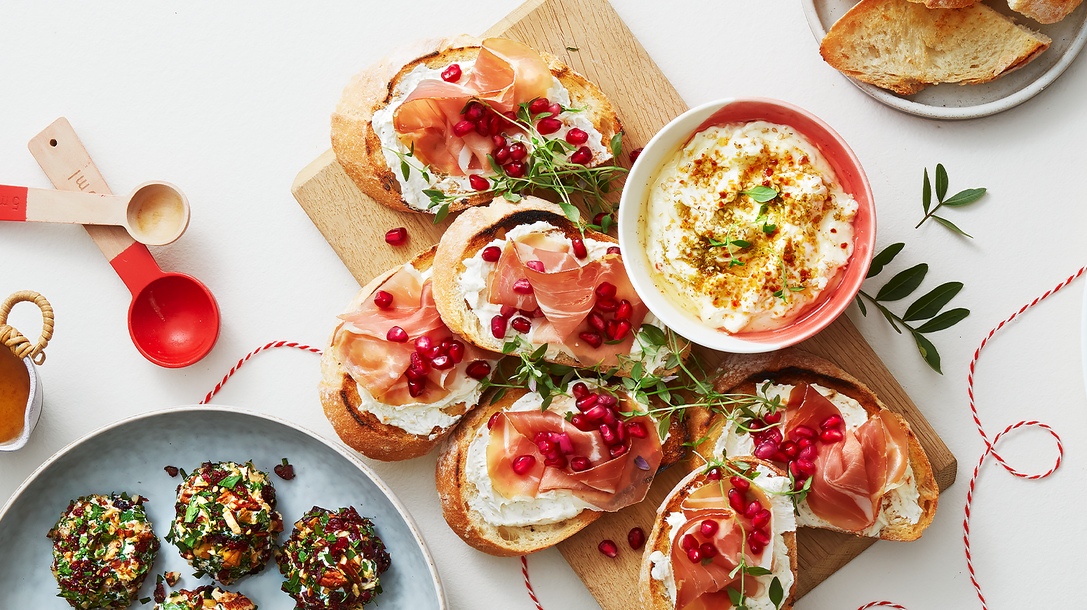 Julete bruschetta med whipped feta, spekeskinke og granateple servert på lys trefjøl på dekket tapasbord. Liten skål med whipped feta på siden.