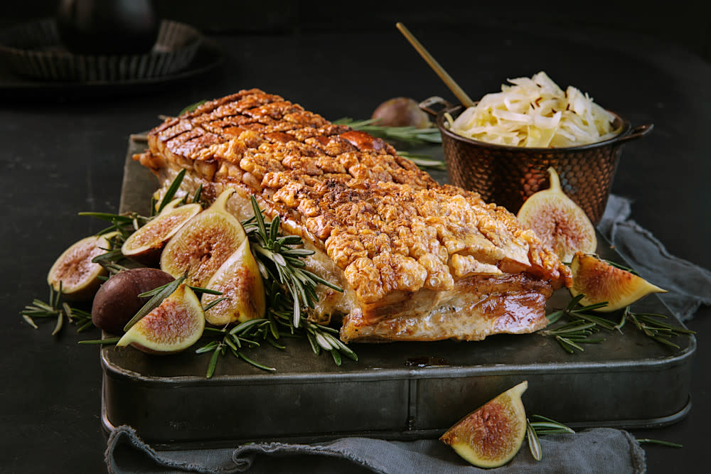 Perfekt ribbe med sprø svor servert på mørk trefjøl med fersk rosmarin og delte ferske fiken rundt. Hjemmelaget surkål i messingskål i bakgrunnen.