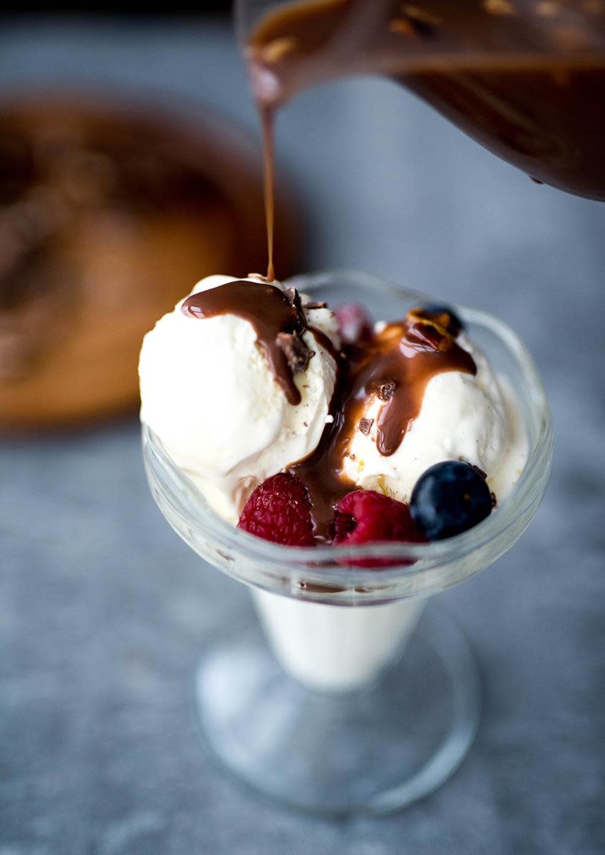 Hjemmelaget Firkløversaus til iskrem, servert over et dessertglass med to kuler vaniljeis, friske bringebær og blåbær. Sausen helles over iskremen fra en glassmugge.