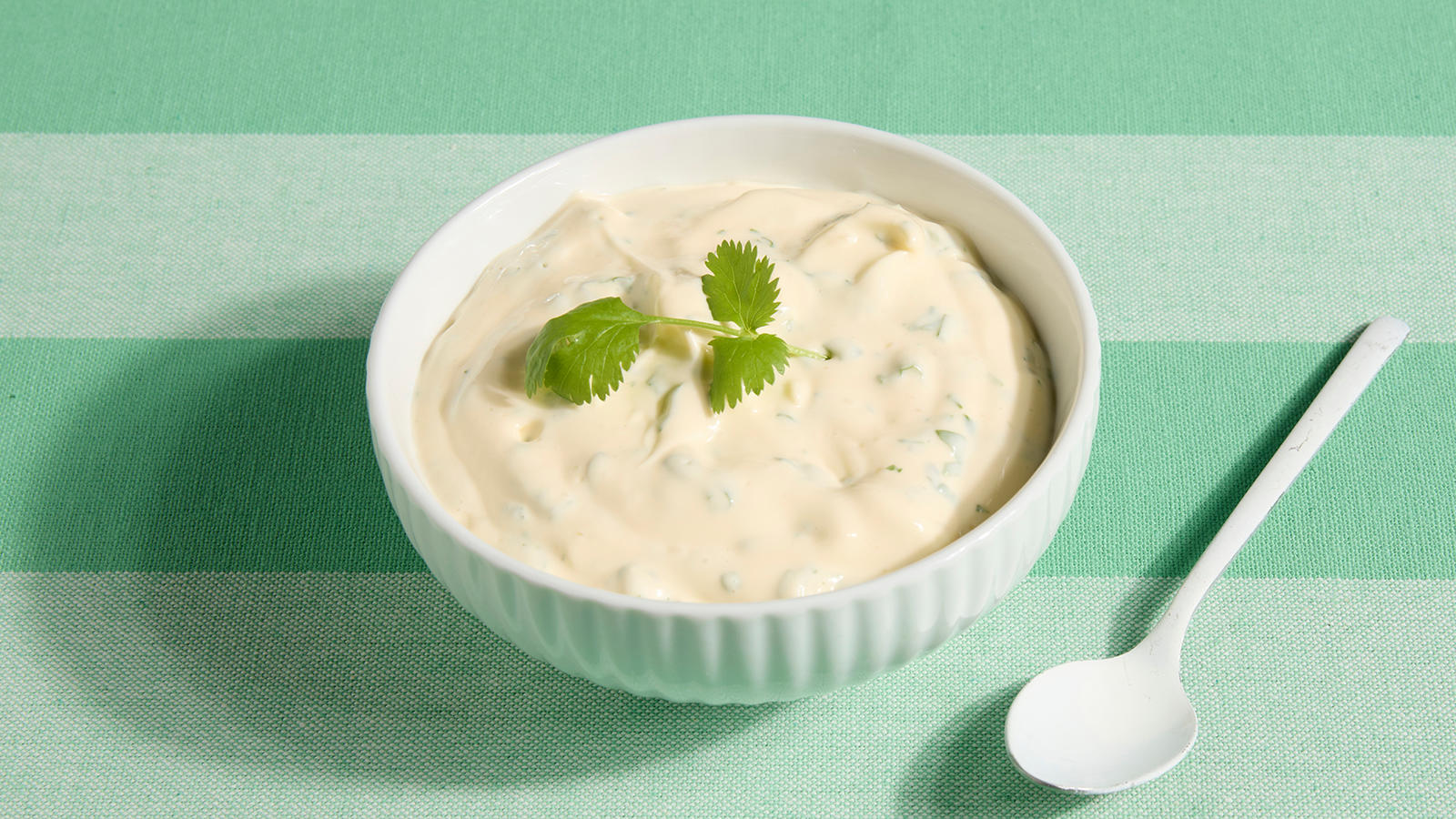 Hjemmelaget aioli med koriander servert i en liten hvit skål på grønnstripete duk. Hvit teskje ved siden av.