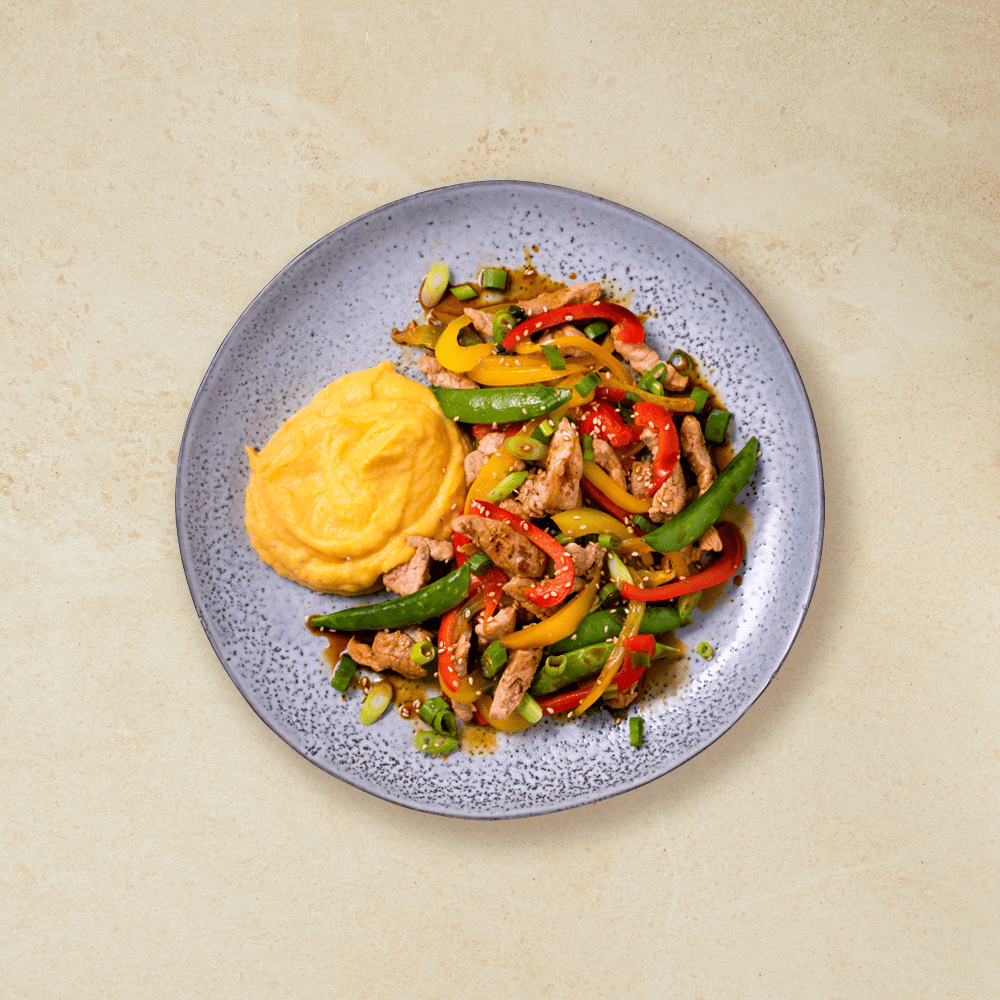 Wok med svinekjøtt og søtpotetmos, paprika, sukkererter og et dryss sesamfrø servert på grå tallerken på beige bakgrunn.