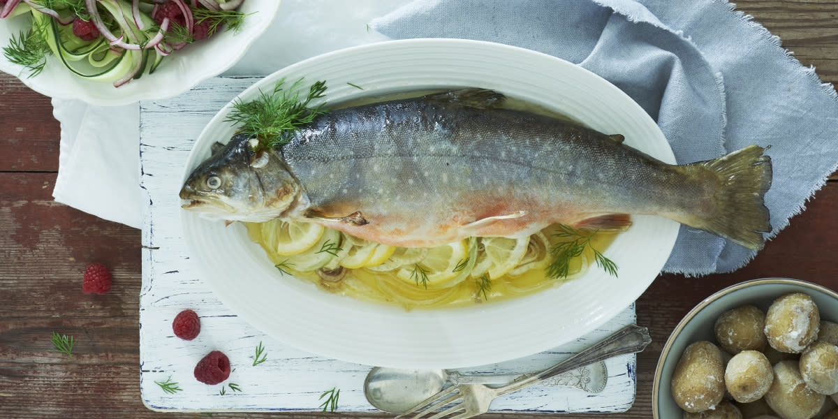 Helstekt ishavsrøye servert på ovalt hvitt fat med sitronskiver. Skåler med saltkokte poteter og agurksalat ved siden av.