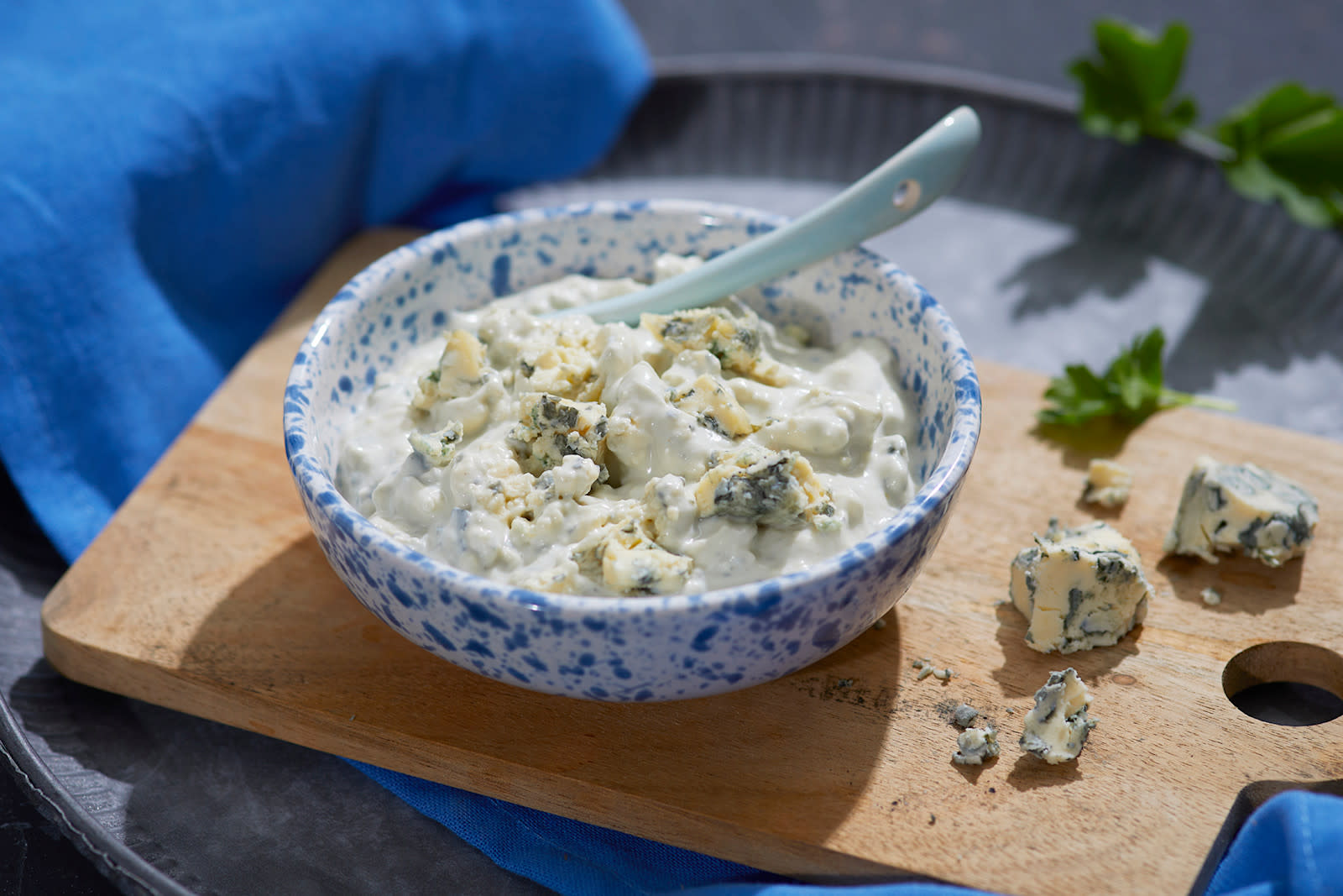 Hjemmelaget blåmuggostdressing servert i blåflekkete hvit skål med smuldret blåmuggost på toppen. Skålen står på en lys liten trefjøl på mørkeblått kjøkkenhåndkle, med blåmuggostsmuler på siden.