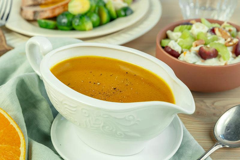 Gylden appelsinsaus i klassisk hvitt sausenebb. Sausen står på et bord sammen med en skål waldorfsalat og en middagstallerken med kalkunstek og rosenkål.