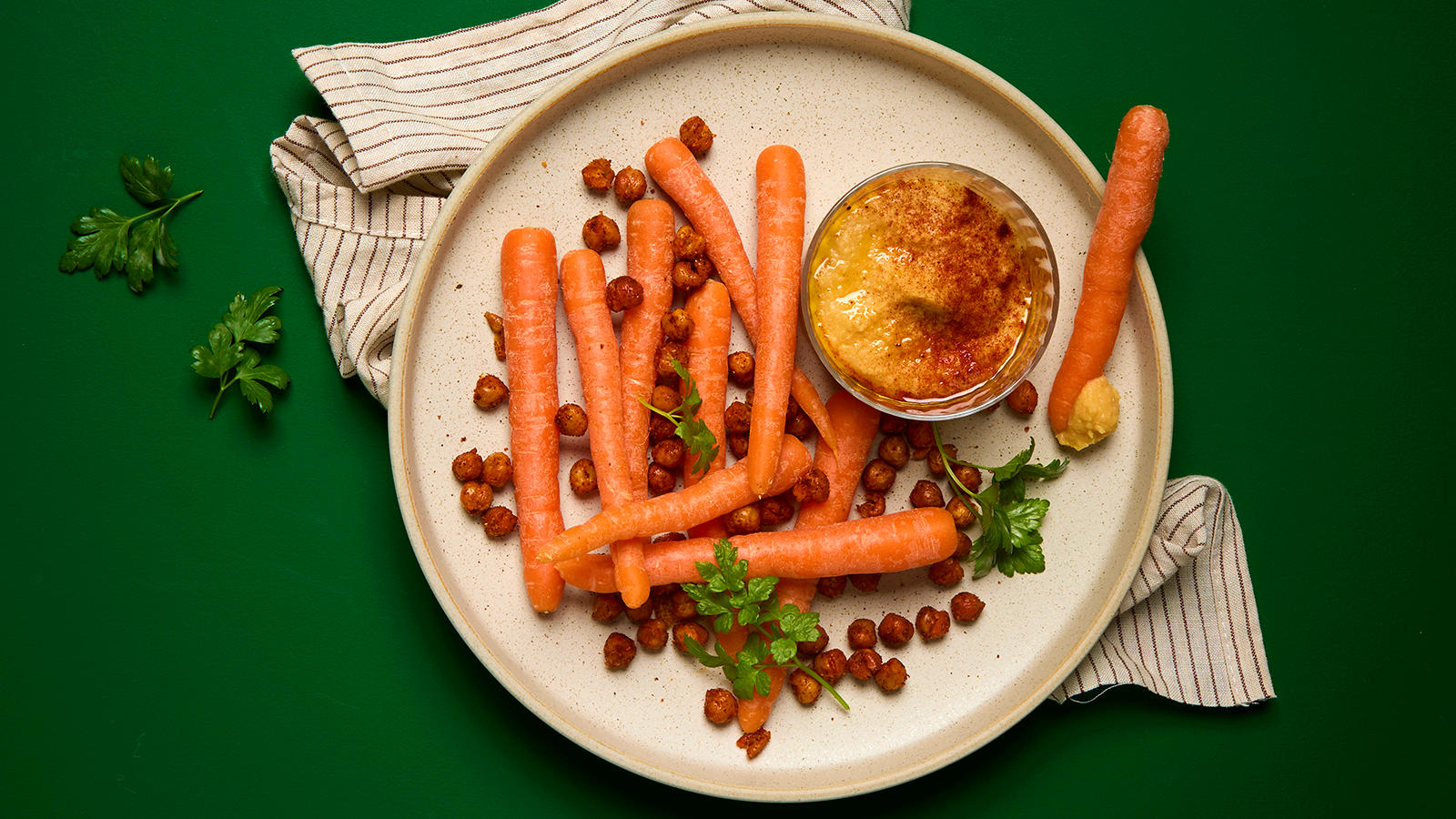 Snackgulrøtter med hummus og sprø kikerter servert på beige fat med frisk persille.