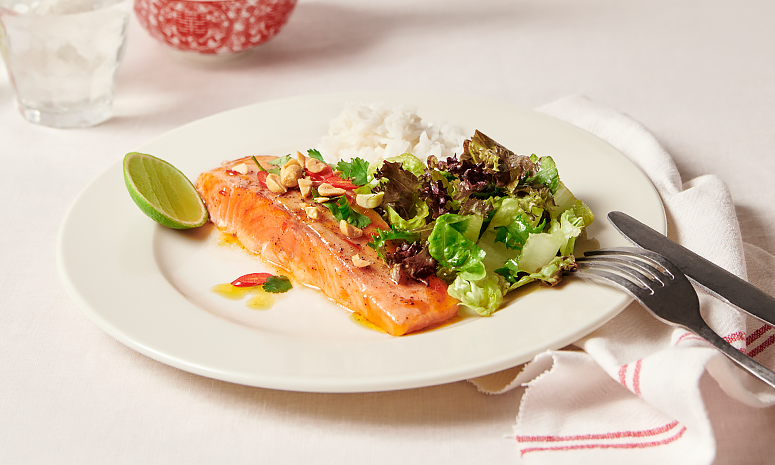 Ovnsbakt sweet chili laks servert på hvit tallerken med ris og salat, toppet med rød chili, hakkede peanøtter og fersk koriander. Liten limebåt ved siden av.