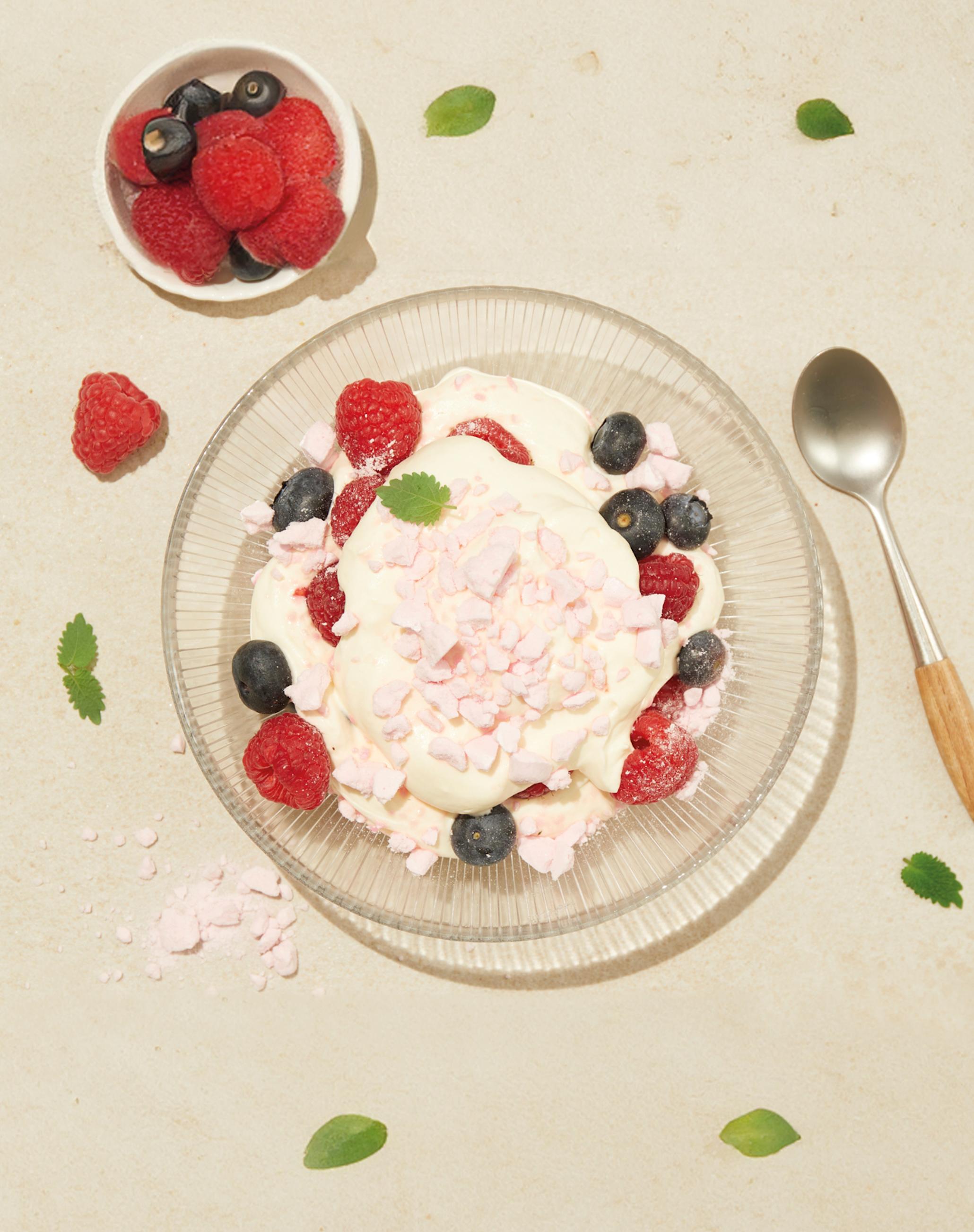 Enkel pavlovadessert à la Eton Mess servert i skål med pisket krem, vaniljekrem og friske bringebær og blåbær. Liten skål med friske bringebær og blåbær ved siden av, og sitronmelisse rundt.