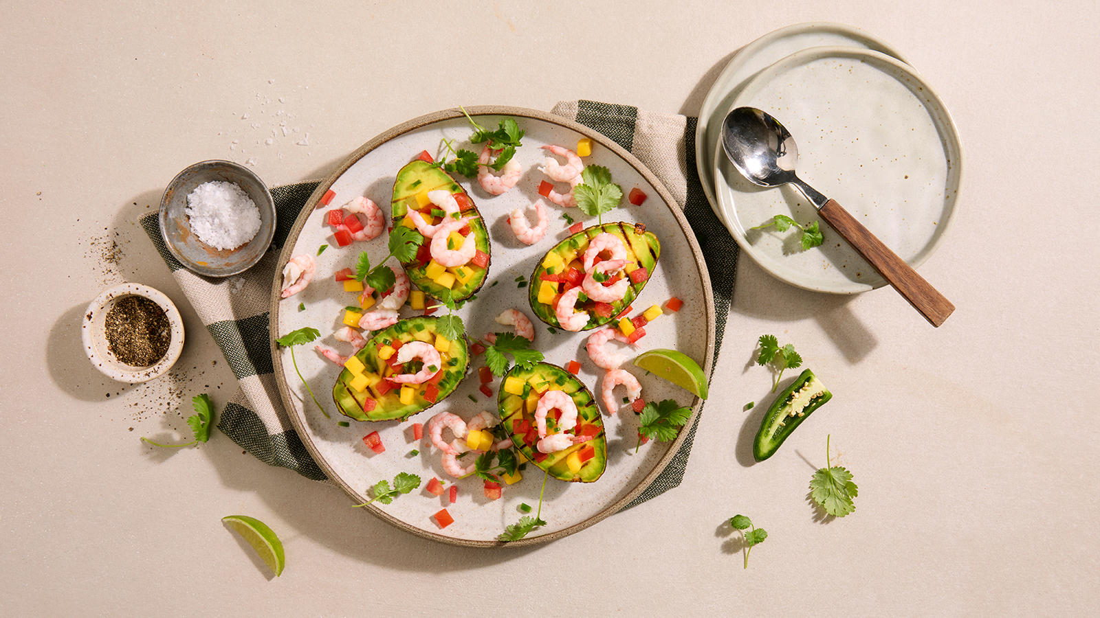 Grillet avokado med mangosalsa og reker, servert på lysegrå tallerken med koriander og limebåter.