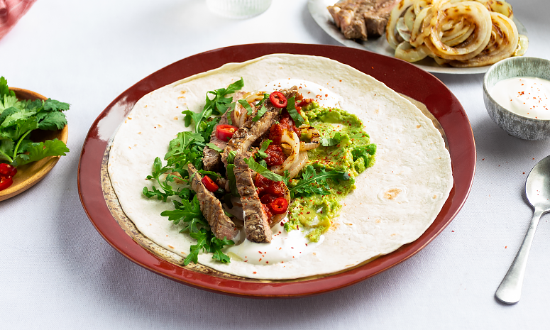 Entrecôte, ruccola, tomat, guacamole, rømme og chili i en tortilla liggende på en rød tallerken. 