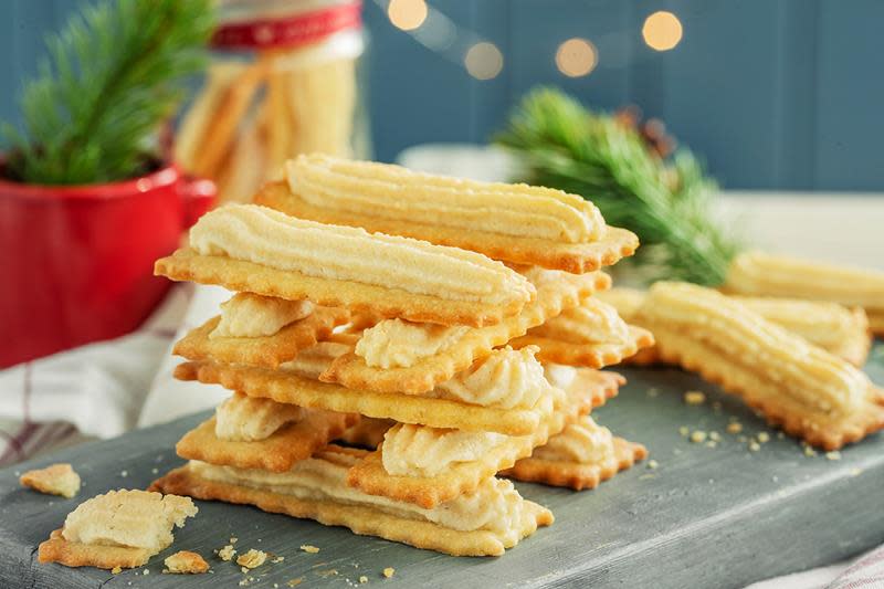 Gylne bordstabelbakkels i stabel servert på mørkegrå trefjøl på julepyntet bord med linduk, rød kopp, julelys og granbar.