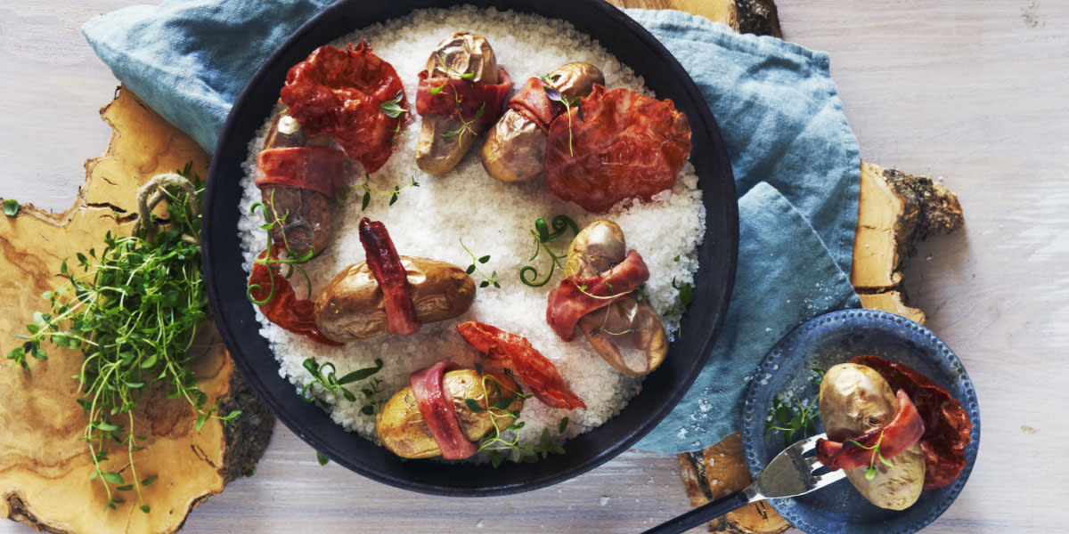 Saltbakte småpoteter med spekeskinke, servert i svart støpejernsform på lyseblå linserviett. Ferske urter, og en liten skål med en potet på gaffel ved siden av.