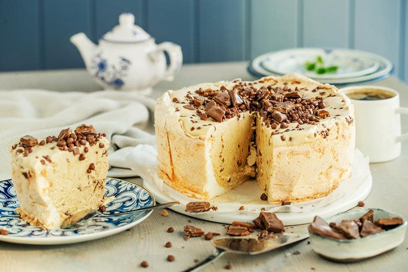 Daimiskake med marengsbunn, servert på hvitt marmorfat. Daimkuler og hakket Daim på toppen. Hyggelig dekket bord, blå bakgrunn.