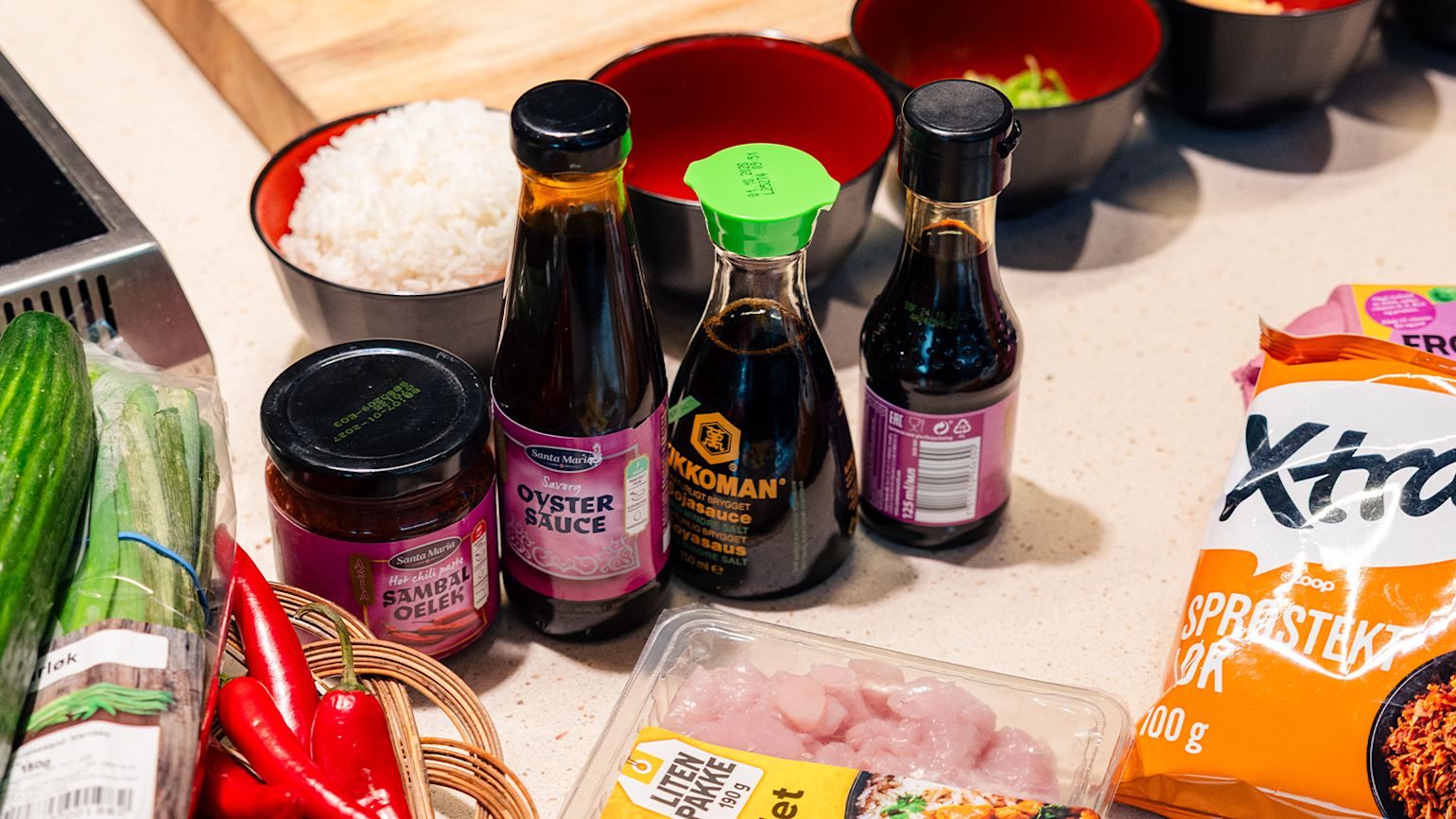 Et nærbilde av ulike asiatiske ingredienser på en kjøkkenbenk, inkludert flasker med østerssaus og soyasaus (Kikkoman), en krukke med Sambal Oelek, rå kyllingbiter, agurk, rød chili, og en pose med sprøstekt løk. I bakgrunnen står to skåler med hvit ris.