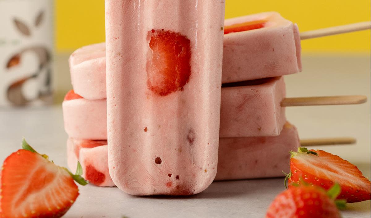 Jordbæris på pinne, med skiver av ferske jordbær i. Pinneisen er stablet oppå hverandre, og det er ferske jordbær rundt.