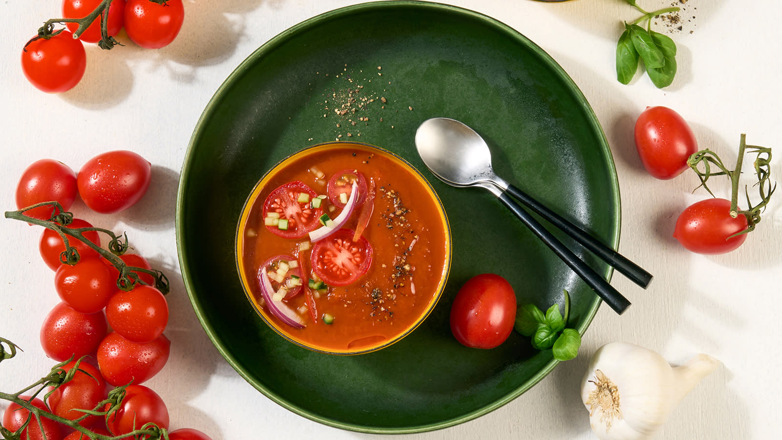 Gazpacho servert i en liten gul skål, med et dryss urter, rødløk, og friske cherrytomater. Friske tomater, hvitløk og basilikum på siden. 