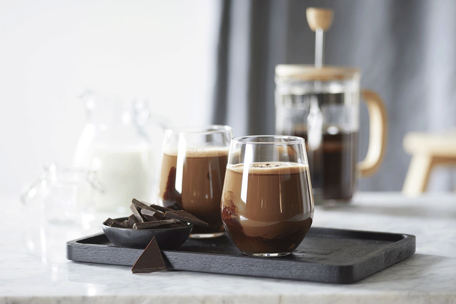 Kaffe mocca i to glass servert på mørk trefjøl med en skål mørk sjokolade i biter ved siden av. Glassmugge med melk, og presskanne med kaffe i bakgrunnen.