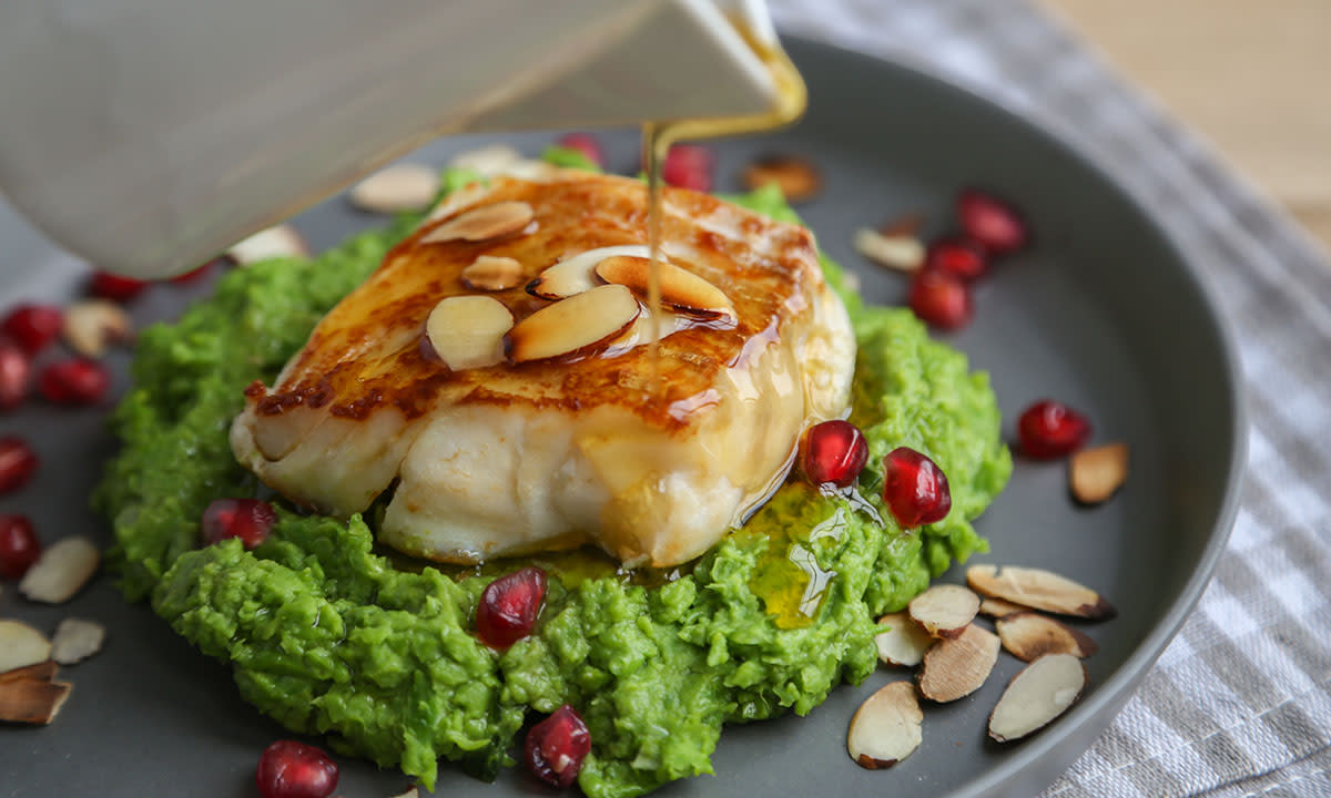 Skrei med nøttesmør og ertestuing servert på grå tallerken med høy kant, og pyntet med skivede mandler og granateplefrø. Smeltet nøttesmør helles over fisken fra en sauseøse.