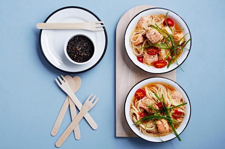 Rød curry med laks og nudler servert i to hvite boller på lys trefjøl, med tomater og finsnittet vårløk på toppen. Lys blå bakgrunn.