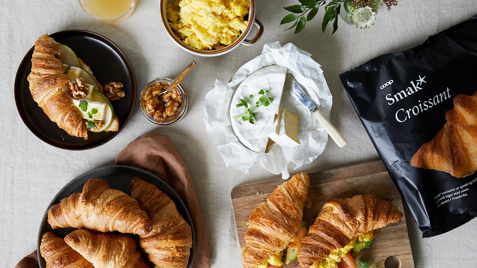 Smak* croissanter servert med to forskjellige fyll på dekket frokostbord med juice, eggerøre, brie og en pose Smak* croissanter.