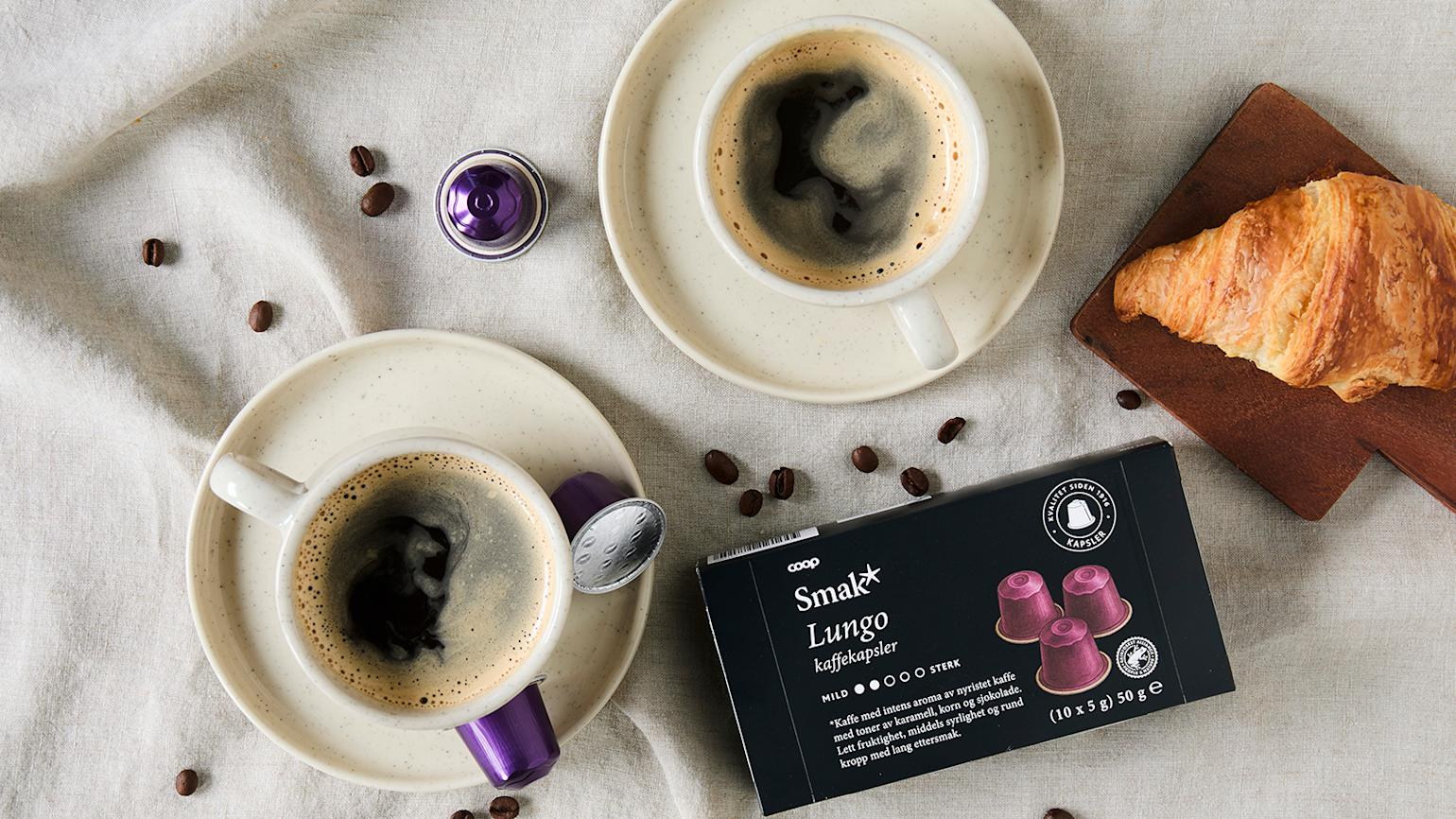 Smak Lungo kaffekapsler på linduk med to kaffekopper, hele kaffebønner og en croissant.