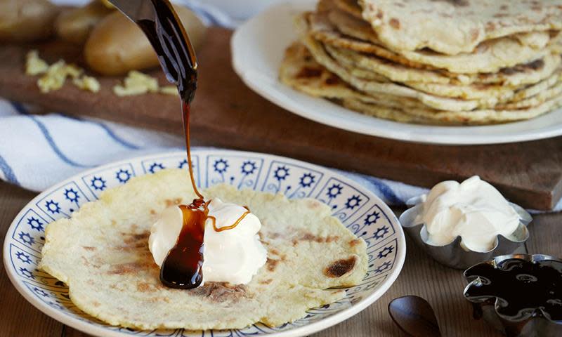 Hjemmelagde potetkaker av potetrester servert på blåmønstret tallerken, på trebord. Stabel med potetkaker i bakgrunnen. Potetkaken på tallerkenen har en klatt rømme på toppen, med ringlet mørk sirup over.