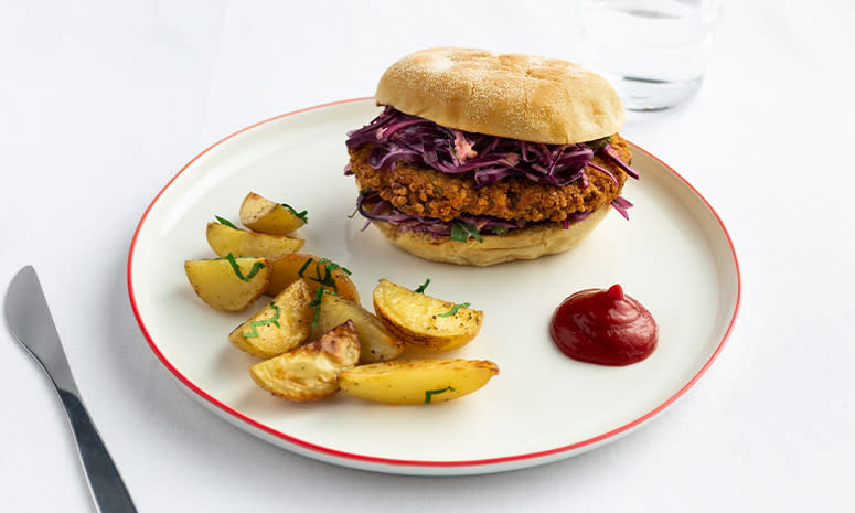 Vegetarburger med rødkålslaw, potetbåter og en dæsj ketchup ligger på en hvit tallerken med rød kant, det ligger en kniv til venstre for tallerkenen