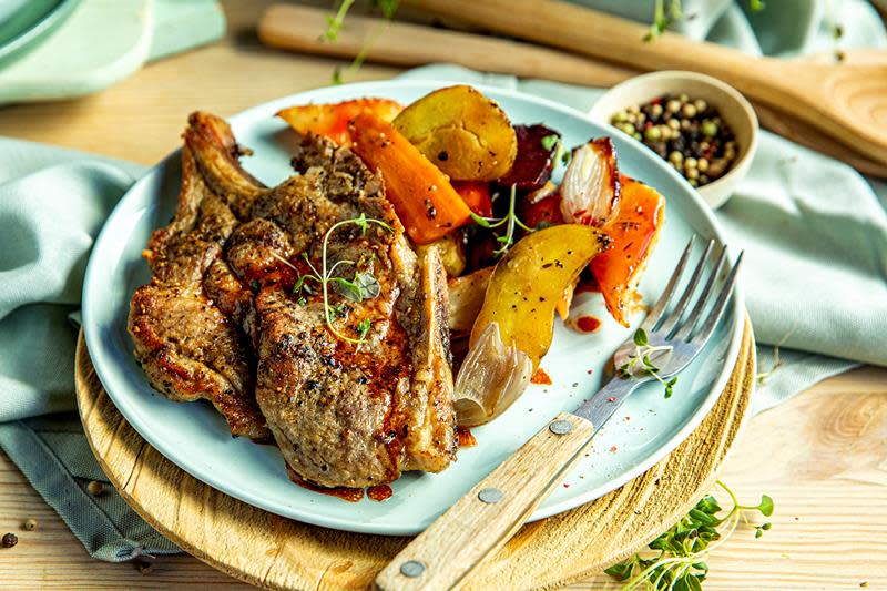 Saftige nakkekoteletter med ovnsbakte rotgrønnsaker i høstlige farger, servert på lyseblå asjett på dekketallerken i tre. Tallerkenen står på trebord med lyseblått kjøkkenhåndkle og en liten skål hel pepper.