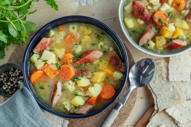 Lys lapskaus med kjøttpølse og rotgrønnsaker, servert i mørk suppeskål på lyst bord. Flatbrød, hel pepper og ferske urter ved siden av.