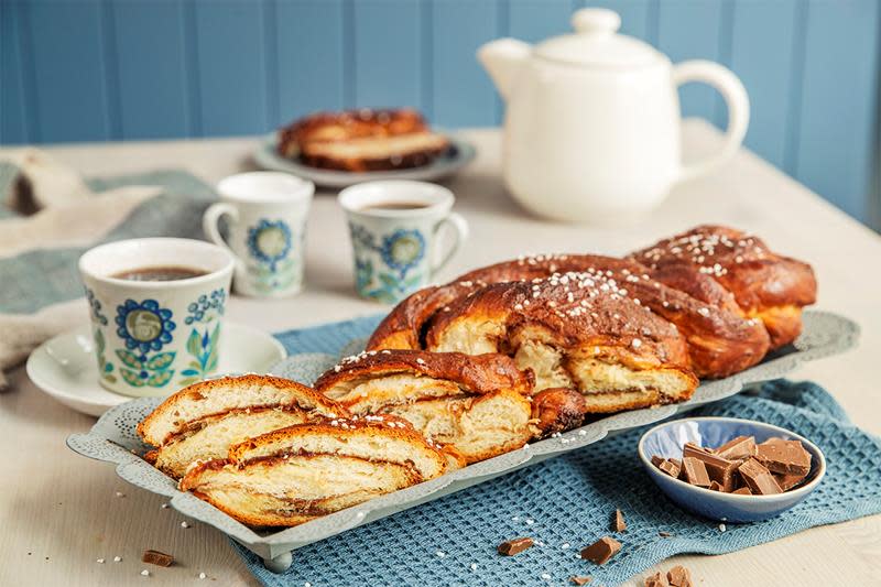 Avlang kringle med sjokoladefyll servert på hyggelig dekket kaffebord, med blå heklet serviett under. Liten skål med sjokoladebiter ved siden av, og skiver av kringle på en asjett.