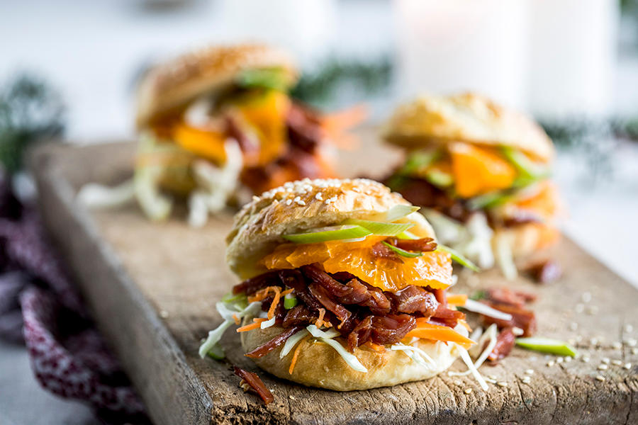 Pinnekjøttsliders; miniburgere med revet pinnekjøtt, finsnittede grønnsaker og klementinskiver servert på rustikk trefjøl med granbar i bakgrunnen.