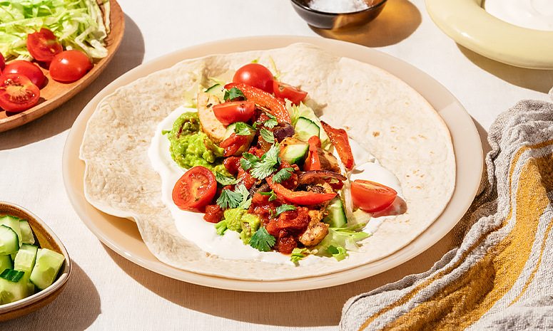 Wrap med kylling, salat og grønnsaker på en beige tallerken med grønnsaker på et fat i bakgrunnen, og et gult håndkle på høyre side av tallerkenen