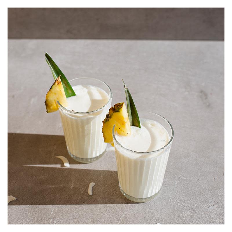 To glass med hvit Piña colada mocktail, toppet med fersk ananas og pyntet med grønt ananasblad. Grå bakgrunn.