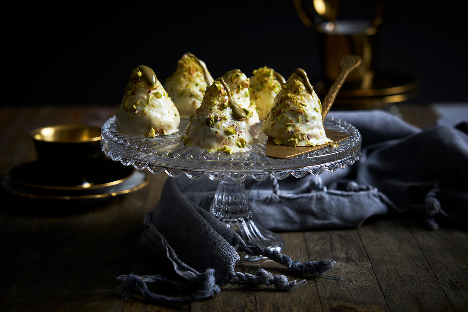 Kokosboller med pistasjekrem servert på stettfat i glass, på mørkt trebord med kaffeservise i bakgrunnen.