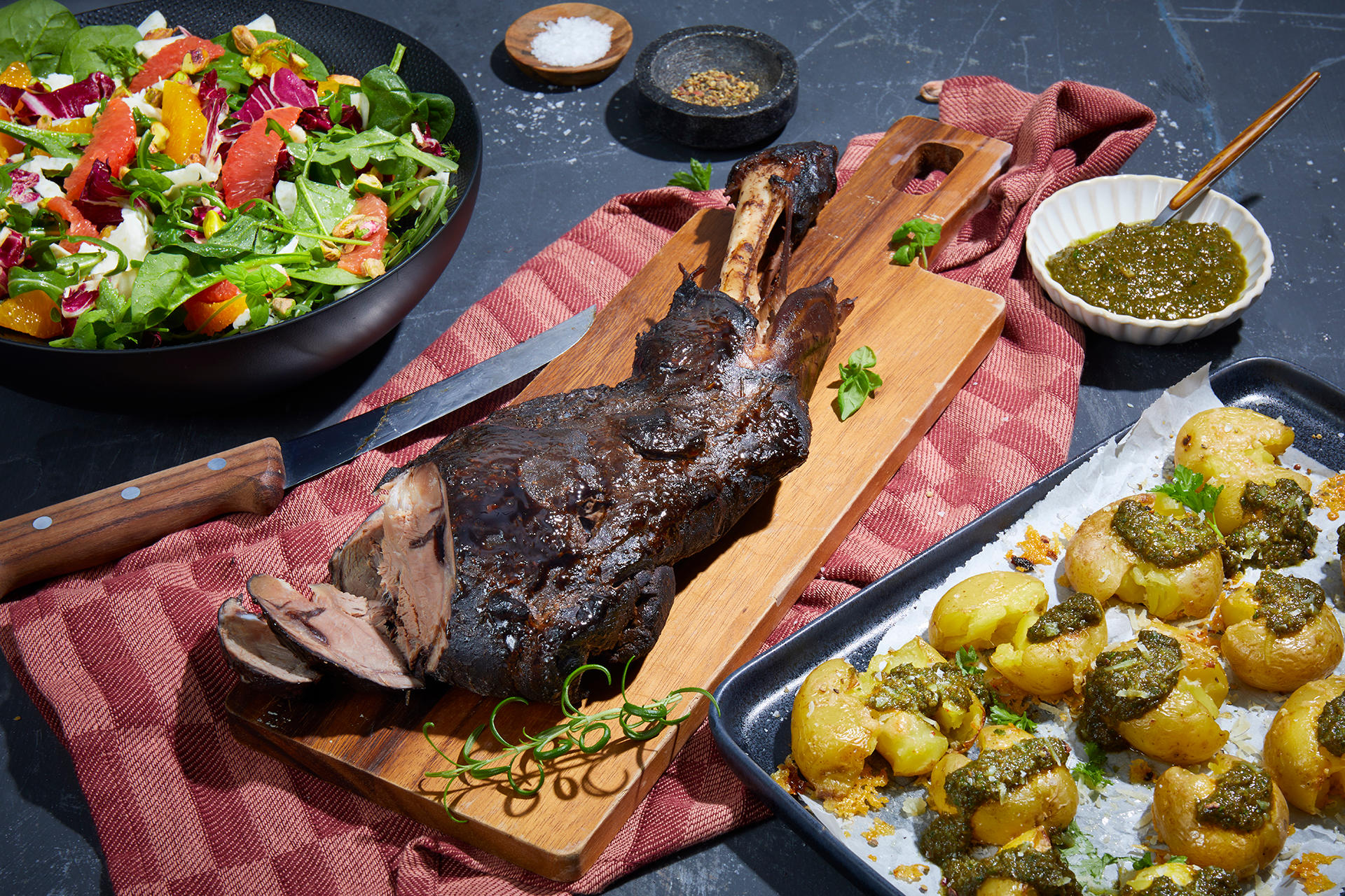 Rødvinsmarinert lammebog med appelsin- og fennikelsalat og parmesanpoteter med urtepesto servert på trefjøl på mørk bakgrunn. Parmesanpoteter på fat til høyre, og salat i mørk bolle til venstre. Små skåler med urtepesto, salt og pepper i bakgrunnen.