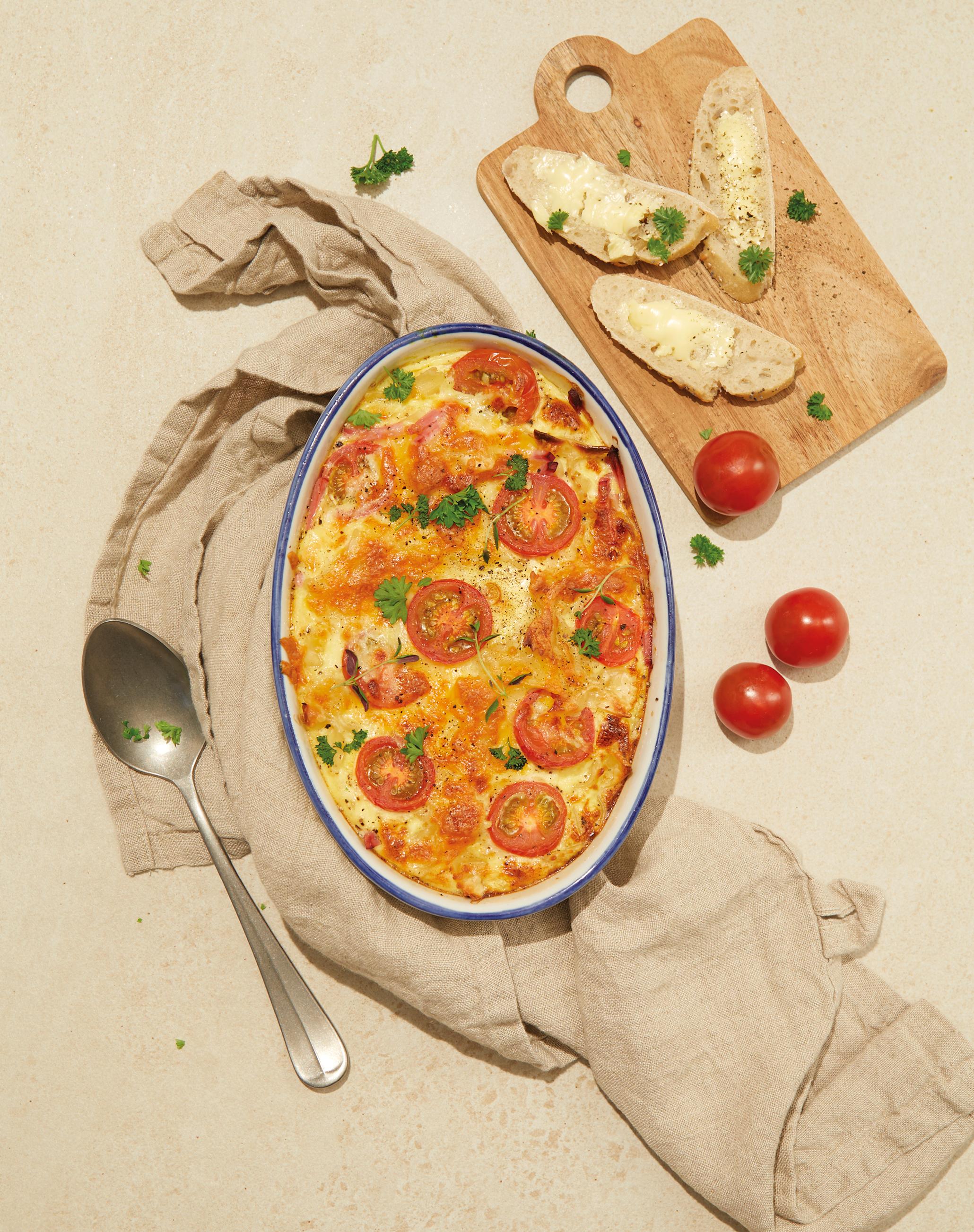 Ovnsbakt omelett med pasta, skinke og tomater, servert i oval ildfast form på beige bakgrunn med linhåndkle under. Liten trefjøl med smurte brødskiver ved siden av.