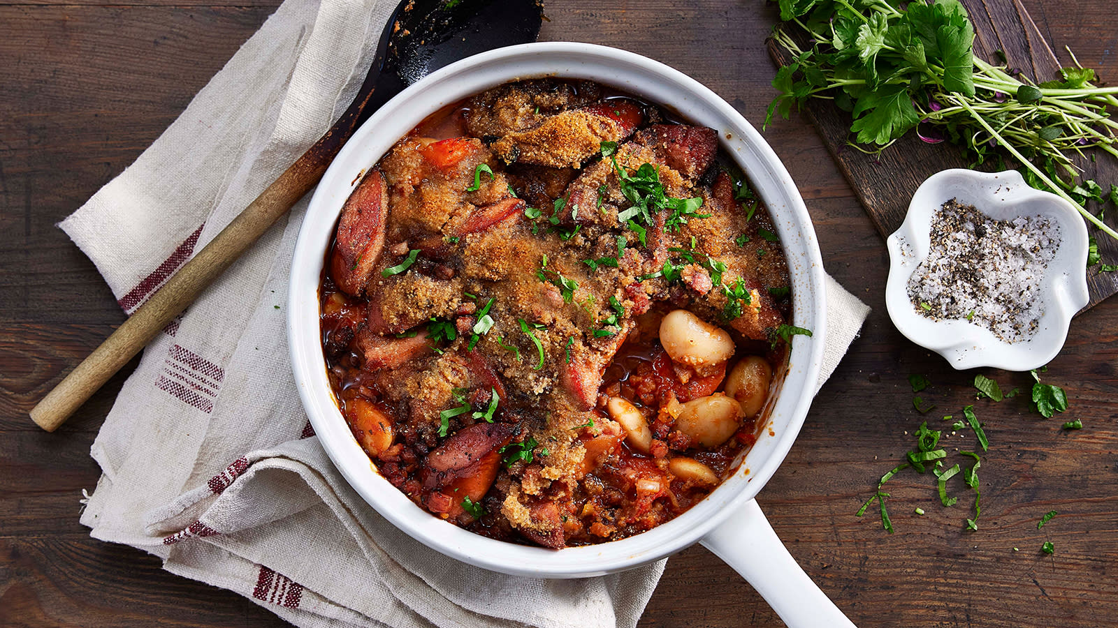 Cassoulet med kjøttpølser - Fransk bondegryte, servert i hvit kasserolle på rustikk mørkt trebord. Bunt med friske urter og liten skål med salt og pepper ved siden av.