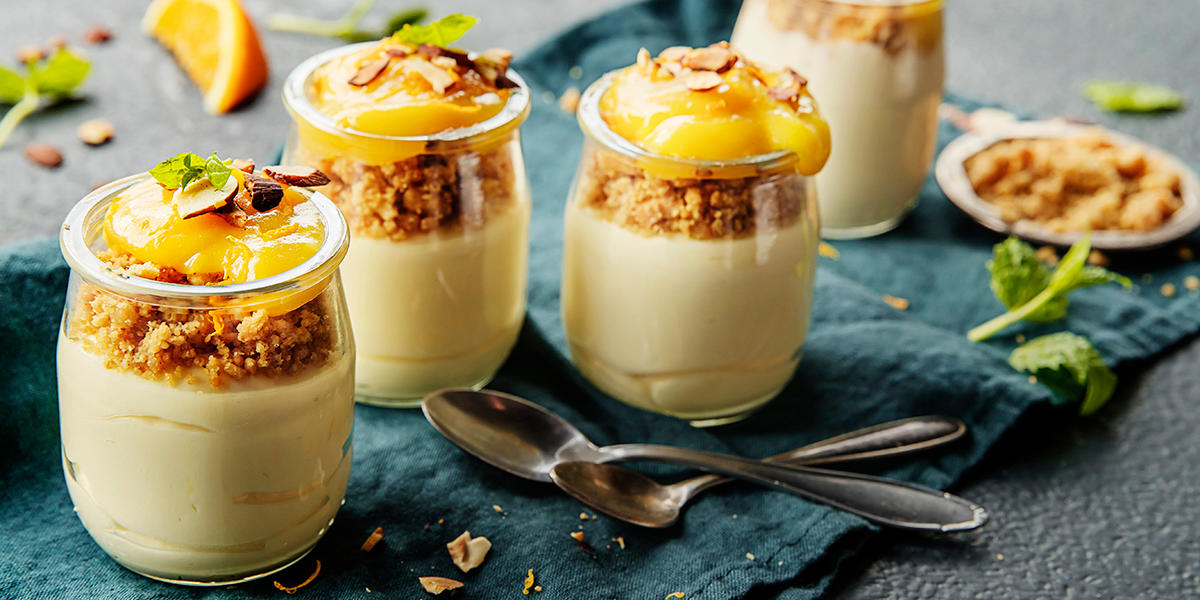 Luftig ostekake i glass med fyldig appelsincurd servert på mørkt bord med blågrønn linserviett. Appelsinskiver, grønne friske urter og en liten skål med knuste kjekssmuler ved siden av.