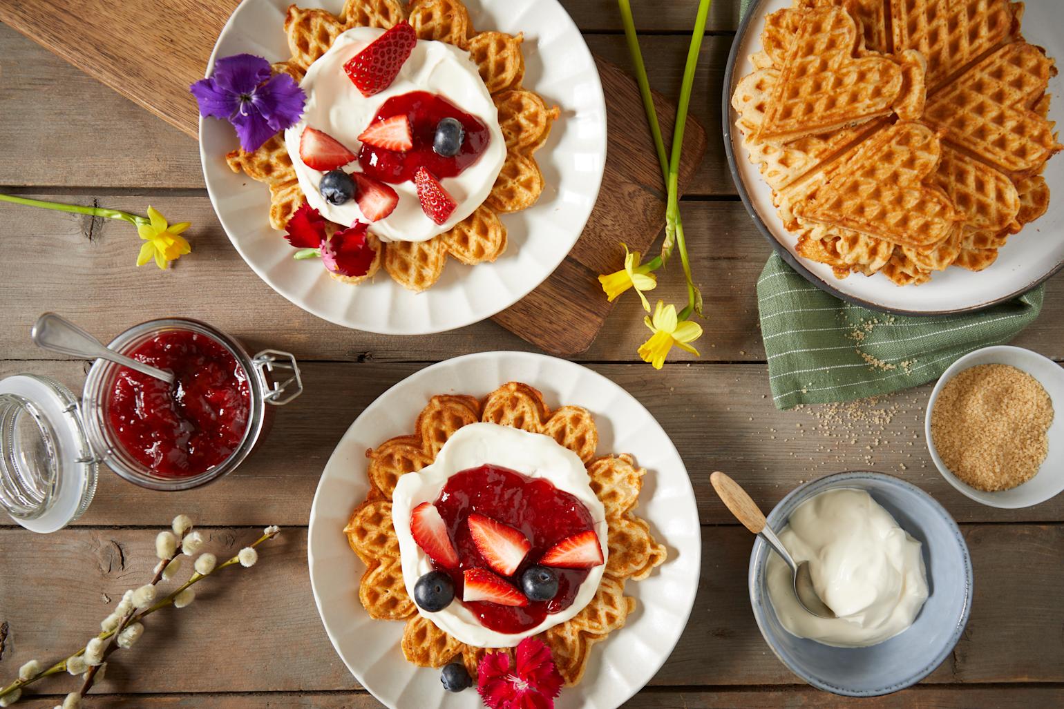 Glutenfrie vafler servert på hvite asjetter med jordbærsyltetøy, friske jordbær og blåbær, pyntet med blomster i forskjellige farger. Asjettene står på et rustikk trebord med påskeliljer, og skåler med tilbehør.