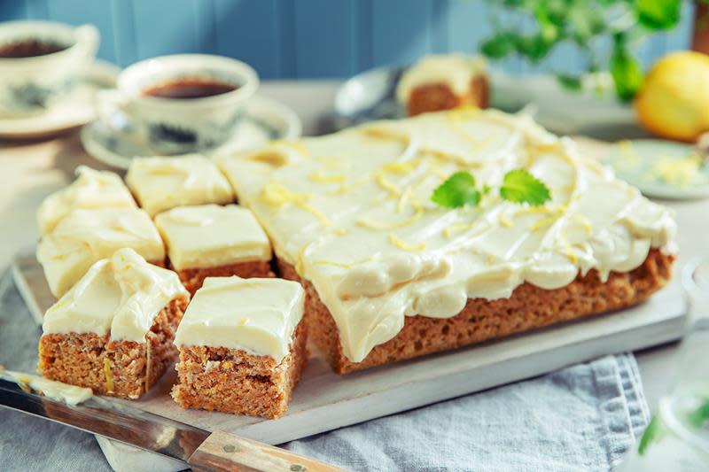 Firkantet gulrotkake i langpanne, delt opp, toppet med ostekrem og pyntet med sitronskall og sitronmelisse. Servert på lys fjøl, på et koselig bord med kaffekopper.