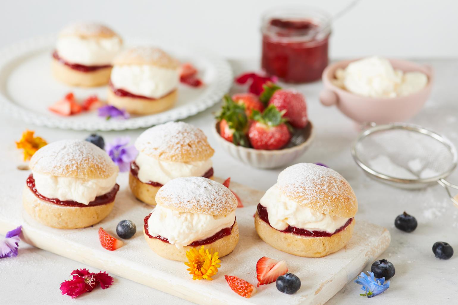 Glutenfrie fastelavnsboller med pisket krem og rødt syltetøy, servert på hvit trefjøl med friske bær og blomster rundt. Sikt med melis, liten rosa skål med pisket krem og liten skål med friske jordbær i bakgrunnen.