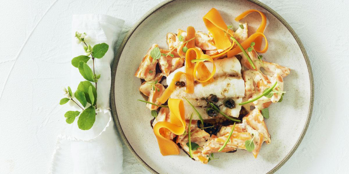 Torsk med chilismør og stuede gulrøtter, grønne urter og gulrøtter i tynne skiver servert på lys tallerken på hvitt bord med hvit serviett ved siden av.