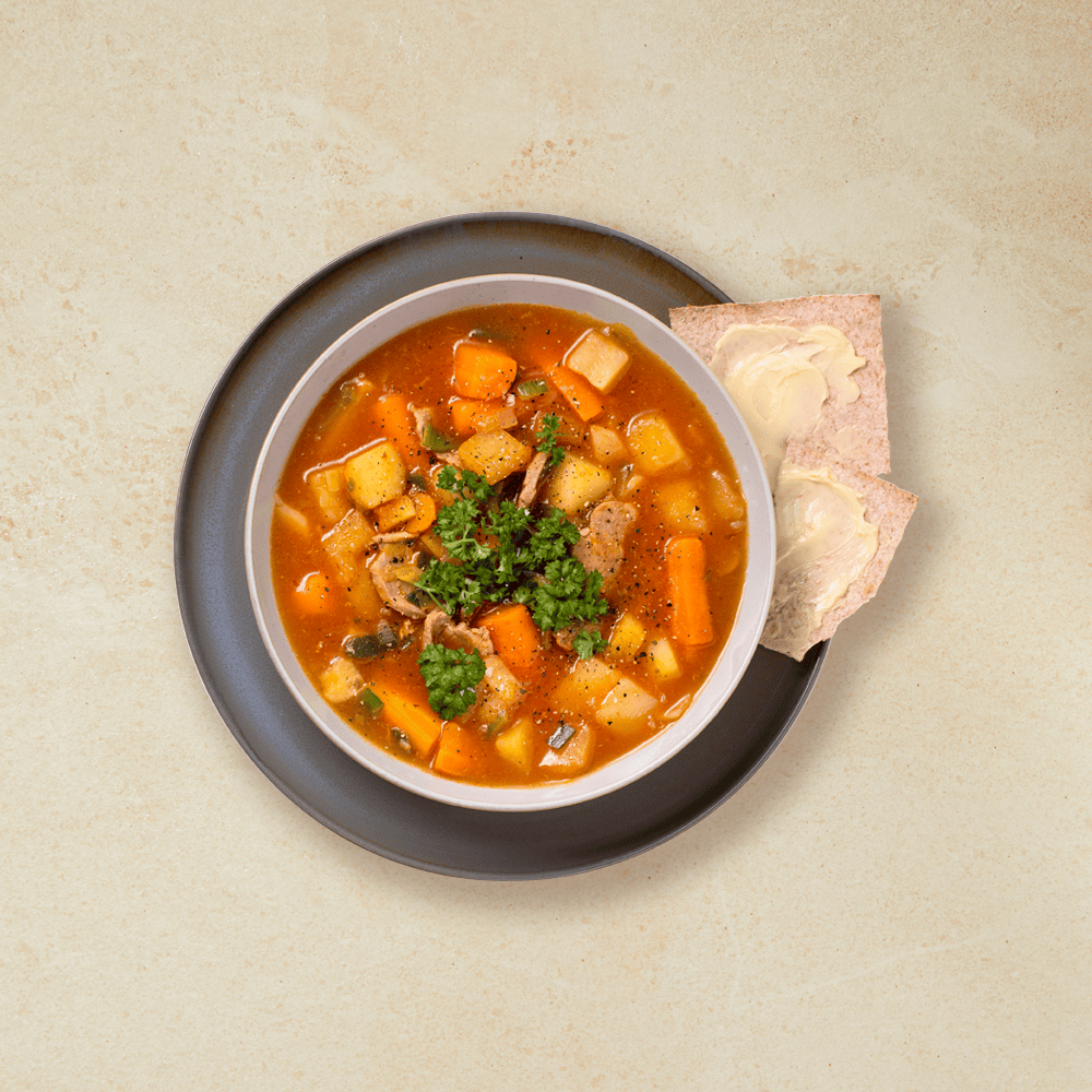 Gryte med svinekjøtt og grønnsaker, servert i hvit skål på mørkegrå asjett med smurt flatbrød på siden. Friske urter drysset over gryta.