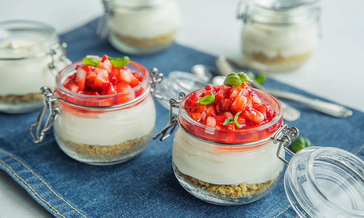 Ostekake i glass med jordbær og basilikum servert på denimserviett med skjeer i bakgrunnen.