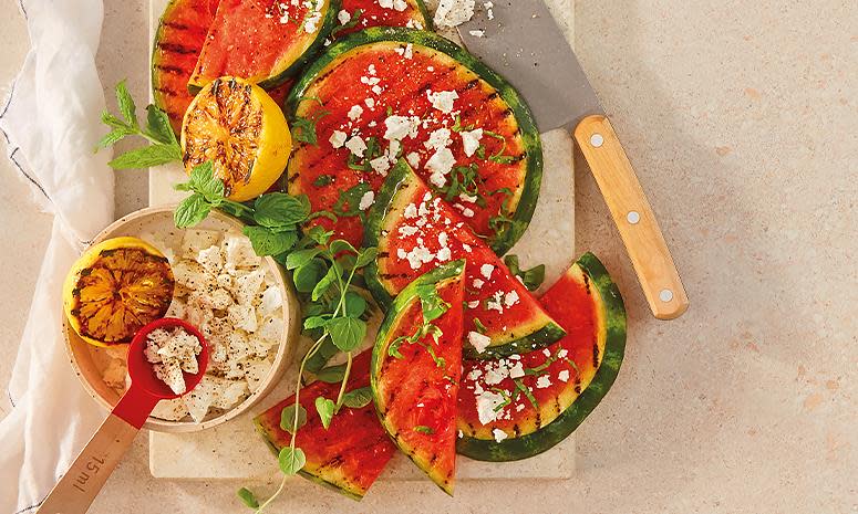 Grillet vannmelon i store runde skiver, med feta smuldret over. Grillet sitron og ferske urter på siden.