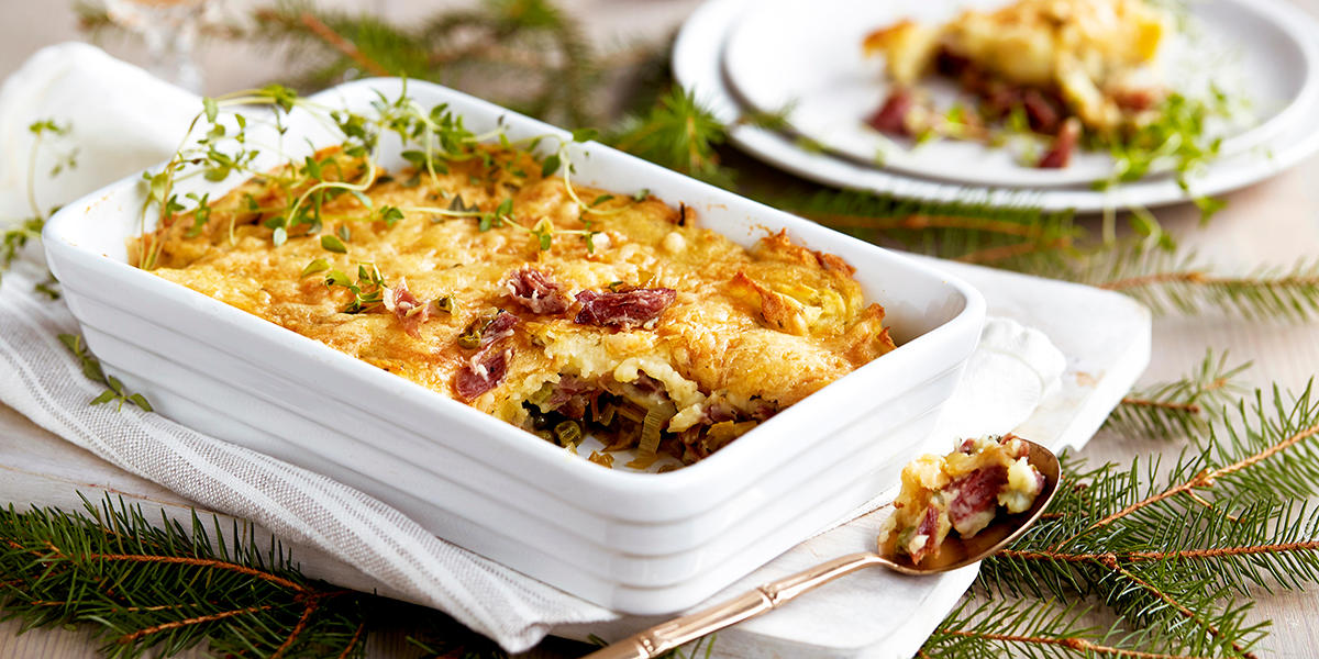 Cottage pie med pinnekjøtt og mandelpotetmos servert i hvit ildfast form, på hvitt fat med granbar under. Servise og bestikk ved siden av.