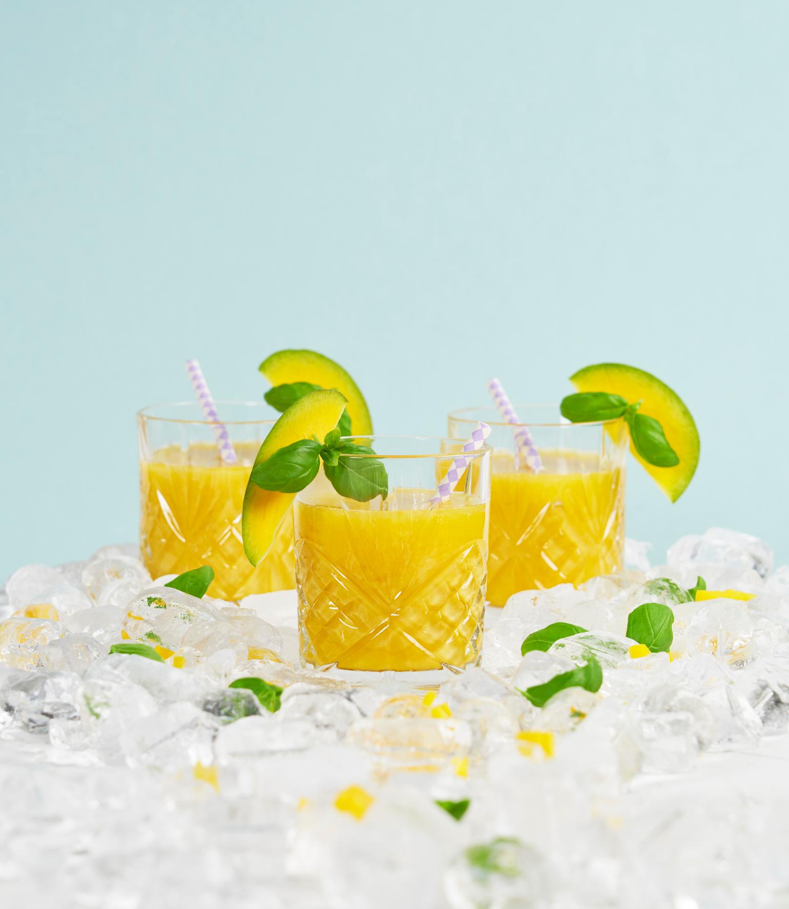 Enkel mango mocktail pyntet med frisk basilikum og skiver av frisk mango, servert i tre mønstrede glass i et hav av isbiter. Blå bakgrunn.