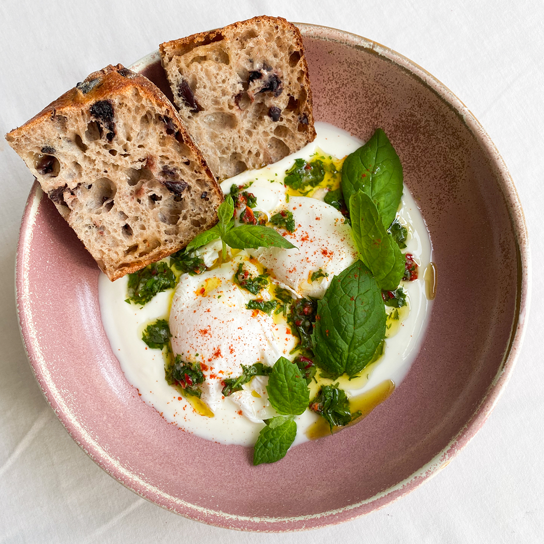 Cilbir - tyrkiske posjerte egg med spicy urteolje, servert i en rosa bolle med yoghurt naturell, urteolje og friske urter, med olivenbrød på siden.