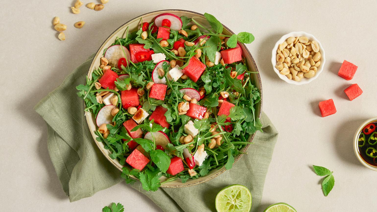 Frisk vannmelonsalat med fetaost og urter servert i lys keramikkskål på grønt kjøkkenhåndkle. Liten skål med peanøtter, og liten skål med dressing på siden.